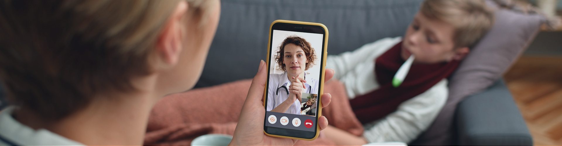 Kvinde holder telefon mens hun snakker med lægen på telefonen og dreng ligger syg i sofaen