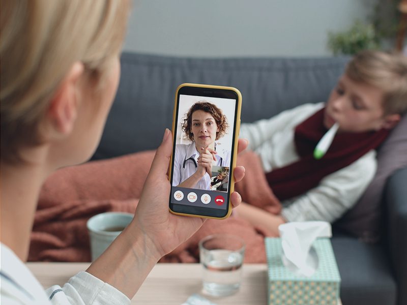 Kvinde holder telefon mens hun snakker med lægen på telefonen og dreng ligger syg i sofaen