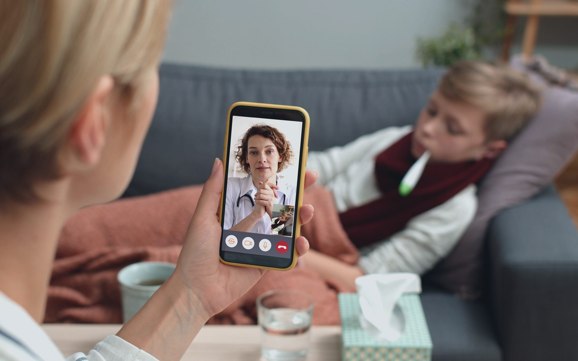 Kvinde holder telefon mens hun snakker med lægen på telefonen og dreng ligger syg i sofaen